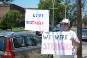 A man holds up a pair of signs saying, "Love the stranger," for "we were strangers." (Photo by Janelle Clausen)